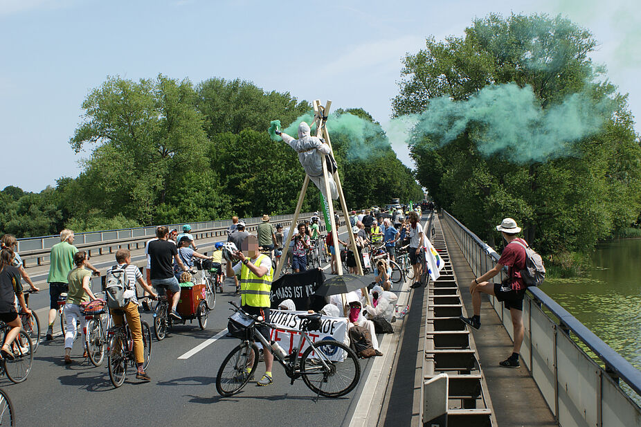 Ende Gelände, Leinemasch bleibt! Aktivisti in weißen Anzügen sitzen mit Bannern unter einem mobilen Tripod, auf dem jemand ein grünes Bengalfeuer abbrennt.